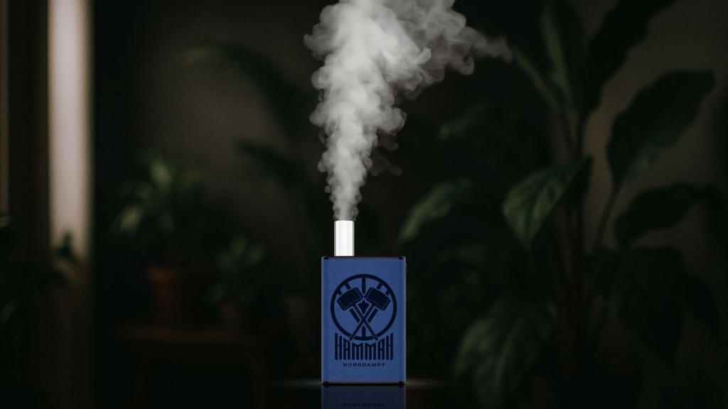 Blauer Vaporizer mit dem Logo „HAMMAH NORDDAMPF“ steht auf reflektierender Oberfläche und stößt eine dichte Dampfwolke aus. Im Hintergrund unscharfe grüne Zimmerpflanzen.